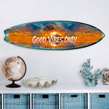 Surfboard-shaped wall art featuring a vibrant orange and blue tropical sunset with silhouetted palm trees and the phrase 'GOOD VIBES ONLY' in white lettering, displayed above a white shelf with decorative objects.