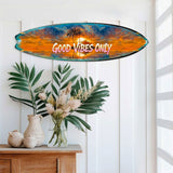 Oval surfboard wall art with a vibrant sunset and silhouetted palm trees, featuring the white text 'Good Vibes Only' in the center, mounted on a white paneled wall above tropical plants.