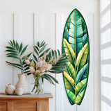 Banana leaf-shaped surfboard wall art featuring layered green and yellow tropical leaves, mounted vertically on a white paneled wall next to a wooden console with leafy plants and textured vases.