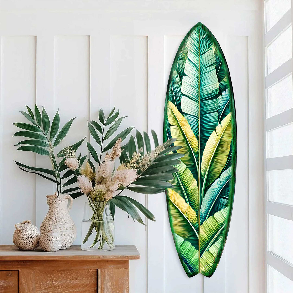 Banana leaf-shaped surfboard wall art featuring layered green and yellow tropical leaves, mounted vertically on a white paneled wall next to a wooden console with leafy plants and textured vases.
