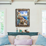 Wall art depicting a coastal village scene seen through rustic, open blue window shutters framed by textured wood, hanging above a teal console in a bright living room.