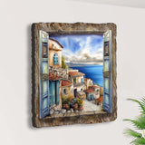 Wall art depicting an open window with blue shutters revealing a coastal town with stone buildings, terracotta roofs, and a calm blue sea under a cloudy sky.