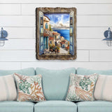 Rustic window frame artwork depicting a sunlit Mediterranean coastal town with terracotta roofs and blue sea under a partly cloudy sky, hung above a light blue sofa with coral-patterned pillows.