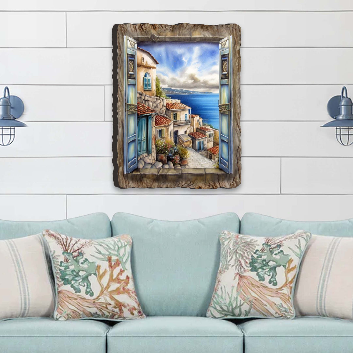 Rustic window frame artwork depicting a sunlit Mediterranean coastal town with terracotta roofs and blue sea under a partly cloudy sky, hung above a light blue sofa with coral-patterned pillows.