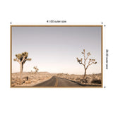 Framed canvas print showing an empty desert road flanked by distinctive Joshua trees under a pale blue sky, with distant low mountains on the horizon.
