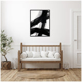 Framed canvas with bold black brushstrokes on a white background, hanging above a wooden bench with white cushions in a bright minimalist room.