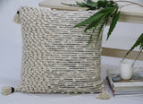 Beige woven throw pillow with textured knots on one side and horizontal woven stripes on the other, adorned with tassels on each corner, placed near books and a white cup.
