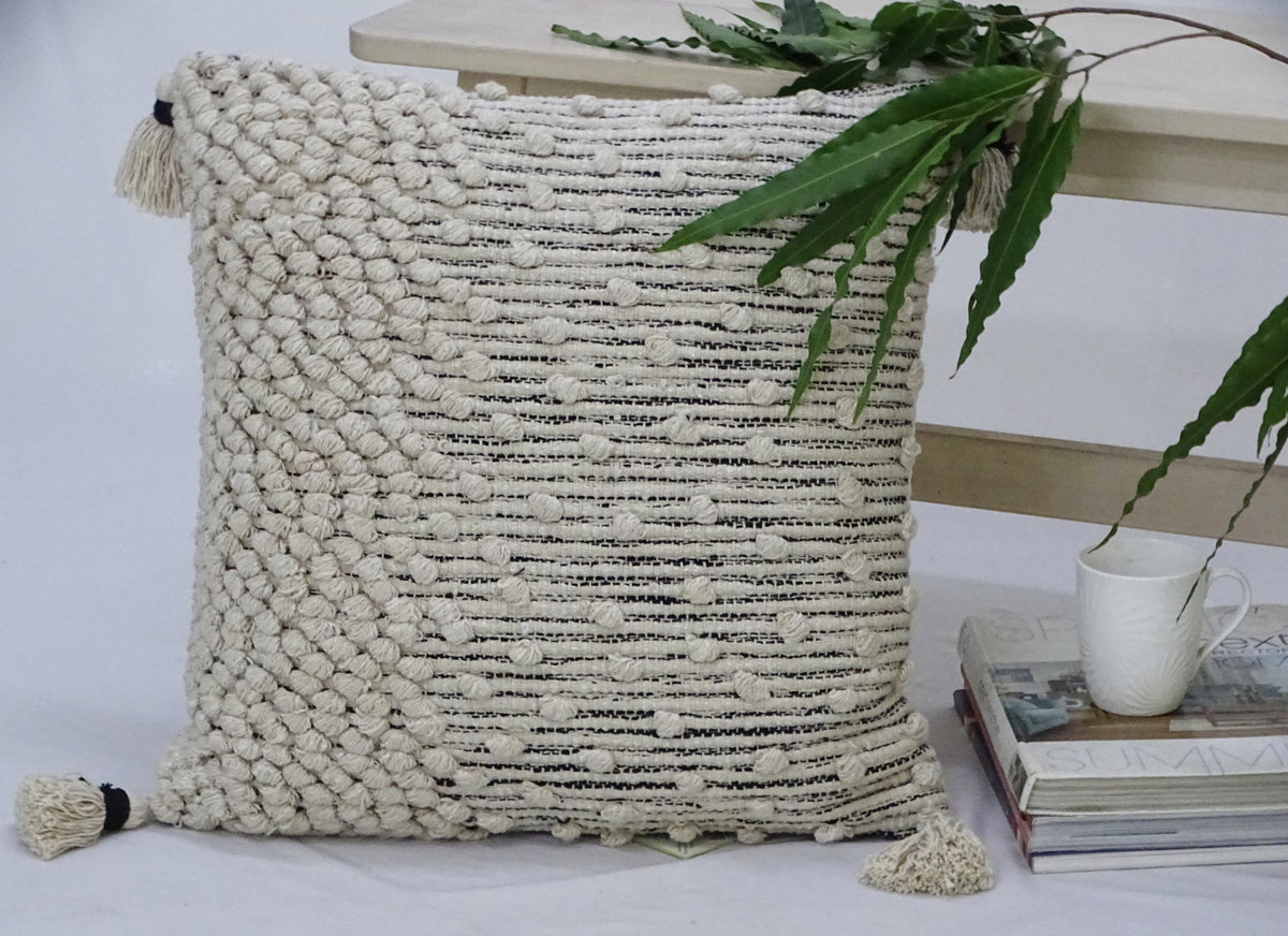 Beige woven throw pillow with textured knots on one side and horizontal woven stripes on the other, adorned with tassels on each corner, placed near books and a white cup.