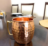 Single hammered copper Moscow Mule mug with brass-colored handle on a dark wooden table, with blurred dining chairs and tableware in the background.