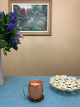 Copper hammered Moscow Mule mug with ice on a blue textured surface, beside an off-white decorative bowl with a floral cutout pattern, under a framed landscape painting.