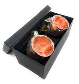 Two copper hammered Moscow Mule mugs with smooth shiny interiors wrapped in plastic, packed side-by-side in a black rectangular gift box with lid.