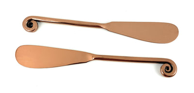 Two copper-finished butter spreaders side by side on a dark surface