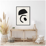 Minimalist living room with a black geometric abstract canvas featuring three black shapes on white wall above a rustic wood console table with decorative pampas grass, lamp, and wicker furniture.