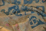 Worn vintage Soumac carpet with muted blue, beige, and pink geometric patterns laid unevenly on a wooden floor, showing faded colors and distressed texture.