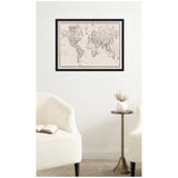 Black-framed vintage style world map with black and white linen texture, hanging on a white wall above two cream upholstered chairs and a brass side table with a white vase.
