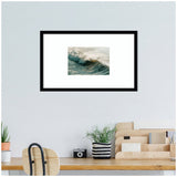 Framed art print of a rolling ocean wave with light reflecting on the water, hanging above a tidy wooden desk with plants and a camera.