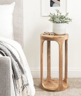 Round Dahl Wood Drink Table with smooth curved legs and a natural wood finish, holding a white textured planter with green foliage and a pair of eyeglasses.