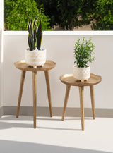 Two round wooden plant stands with three tapered legs, each holding a white ceramic pot with geometric designs and green plants, set against a white railing and green foliage.