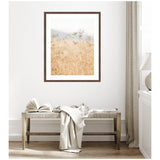 Framed photo of wild fennel plants in golden tones with soft blue-gray sky, hung above a wooden bench draped with a white cloth in a bright room.