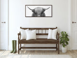 Black and white framed canvas of a Highland cow's head with horns, hung above a rustic wood bench with white pillows, flanked by a pair of green boots and a potted plant.