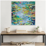 Square framed canvas of an impressionistic water lily pond with green, blue, purple, and pink brushstrokes, hanging above a wooden console table with books, a mug, and a white vase with a trailing plant.