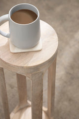 Light wood round stool with a white ceramic mug filled with coffee on a white square coaster, shown from above against a beige textured background.