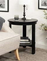 Round black wood side table with a lower shelf holding books, topped with a dark candlestick lamp and a glass, positioned beside a beige sofa with wooden legs.