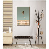 Framed wall art featuring a lone palm tree bent over a pale building under a clear turquoise sky, above a black cushioned bench and a black coat rack with umbrella and hat.