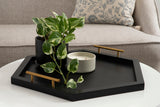 Black hexagon tray with gold handles holding a potted variegated green plant and a white candle on a white round table in front of a beige sofa with a patterned pillow