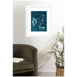 Framed aerial-style art of four humpback whales swimming in deep blue ocean water, hung on a white wall above a dark olive green chair with an open book and a woven basket plant nearby.