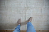 Person wearing beige slip-on shoes and blue jeans standing on a muted beige vintage distressed Oushak carpet with subtle wear patterns.