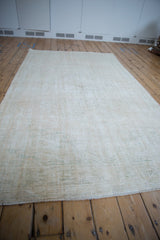 Light beige vintage distressed Oushak carpet with subtle pattern on wooden floorboards in a room with pale walls and baseboard vents.