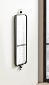 Tall rectangular metal-framed mirror with rounded corners mounted vertically on a white wall, featuring decorative finials at top and bottom, beside a window with black trim.