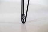 Close-up of a black metal hairpin table leg with an intricate twisted knot detail near the bottom, standing on a neutral floor.