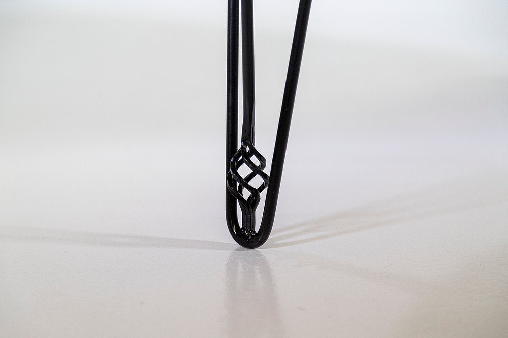 Close-up of a black metal hairpin table leg with an intricate twisted knot detail near the bottom, standing on a neutral floor.