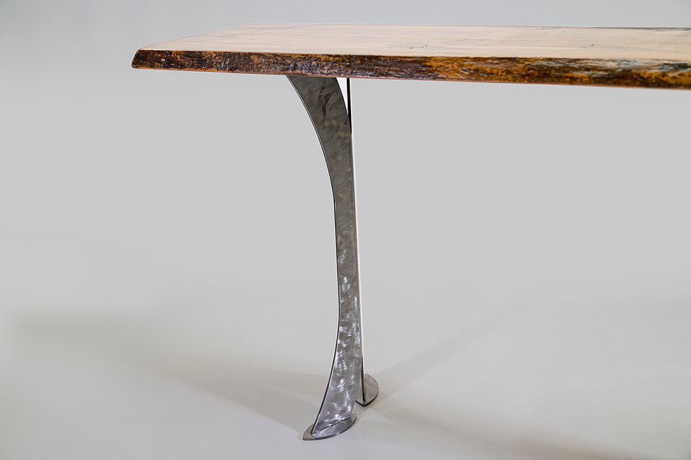 Close-up of a silver curved metal leg with a hammered texture supporting a thick honey-toned wood tabletop with a textured natural edge against a plain light background.