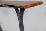 Close-up of a live-edge honey-toned wood tabletop with hammered silver split legs against a plain background