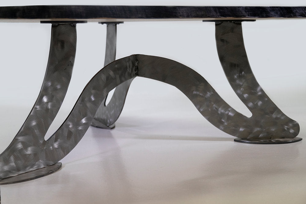 Close-up of a hammered metal table base with flowing, curved scalene-shaped supports in a silver-gray finish under a dark wood tabletop edge.