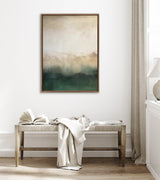 Framed abstract canvas featuring soft gradations of muted beige and green tones suggesting a misty mountain landscape, displayed above a wooden bench with fabric and an open book.