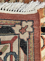 Corner of a hand-knotted rug with a cream and rust floral motif, black and beige geometric patterns, and a cream fringe on a rust border.