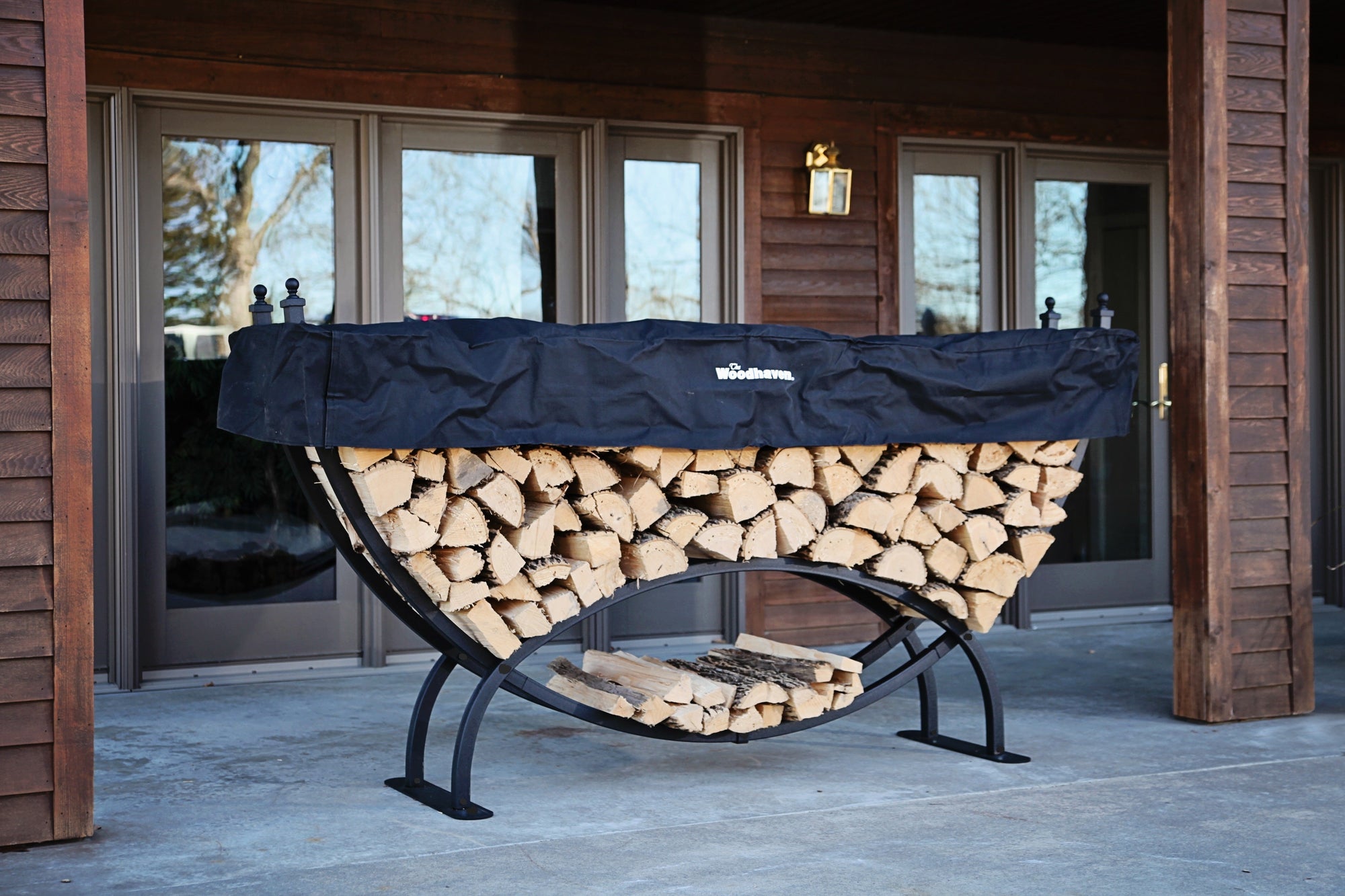 Woodhaven 8 ft Large Crescent Firewood Rack with seasoning cover holding firewood outside a house