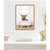 Brown Highland cow with long horns and shaggy hair sitting in a white vintage clawfoot bathtub against a white paneled wall with wooden floor, framed in light wood.