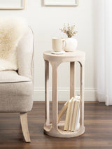 Light wood Dahl Wood Drink Table with open circular design and lower shelf holding books, placed beside a beige upholstered chair with a white fur throw on dark wood floor