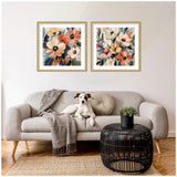 Two framed abstract floral prints with warm orange, yellow, and blue tones hang above a light gray sofa with a white and brown dog resting on it, next to a black woven round coffee table with a small plant.