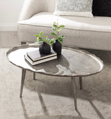 Oval metal coffee table with a scalloped edge and textured surface, holding two black vases with green plants and two stacked books, in front of a light gray sofa.