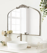 Tall arched bathroom mirror with ornate floral embellishment at the top, reflecting a window, above a white oval vessel sink and sleek modern faucet on a light countertop.