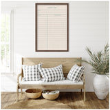 Wooden bench with white cushioned seat and three gray and white buffalo check pillows under a framed large vintage-style library card on white shiplap wall.