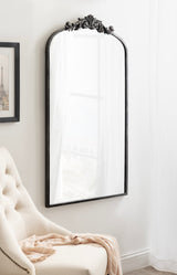 Tall arched wall mirror with ornate black decorative crest at top, hanging above a beige tufted chair with a light pink throw draped over it, in a softly lit room.