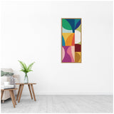 Abstract geometric art with green, blue, orange, pink, and purple shapes arranged vertically in a wooden frame on a white wall above a wooden side table with a glass vase and green fern.