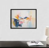 Abstract coastal-inspired painting with soft blues, pinks, and oranges blending into white, framed in black and hung on a light gray wall above a black desk.
